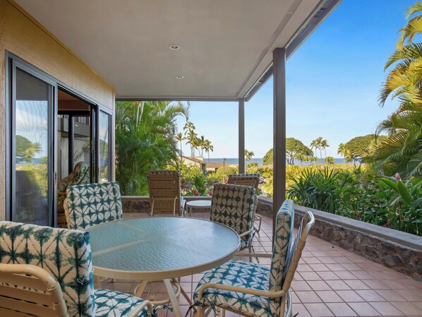 A covered outdoor seating area with a round table and chairs, surrounded by tropical plants and a view of the ocean.