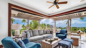 A spacious living area with a ceiling fan, a large sofa, armchairs, and a coffee table. The room has large windows offering a view of palm trees and the ocean. - Recently Renovated & Professionally Managed Ocean View Villa (Wailea)