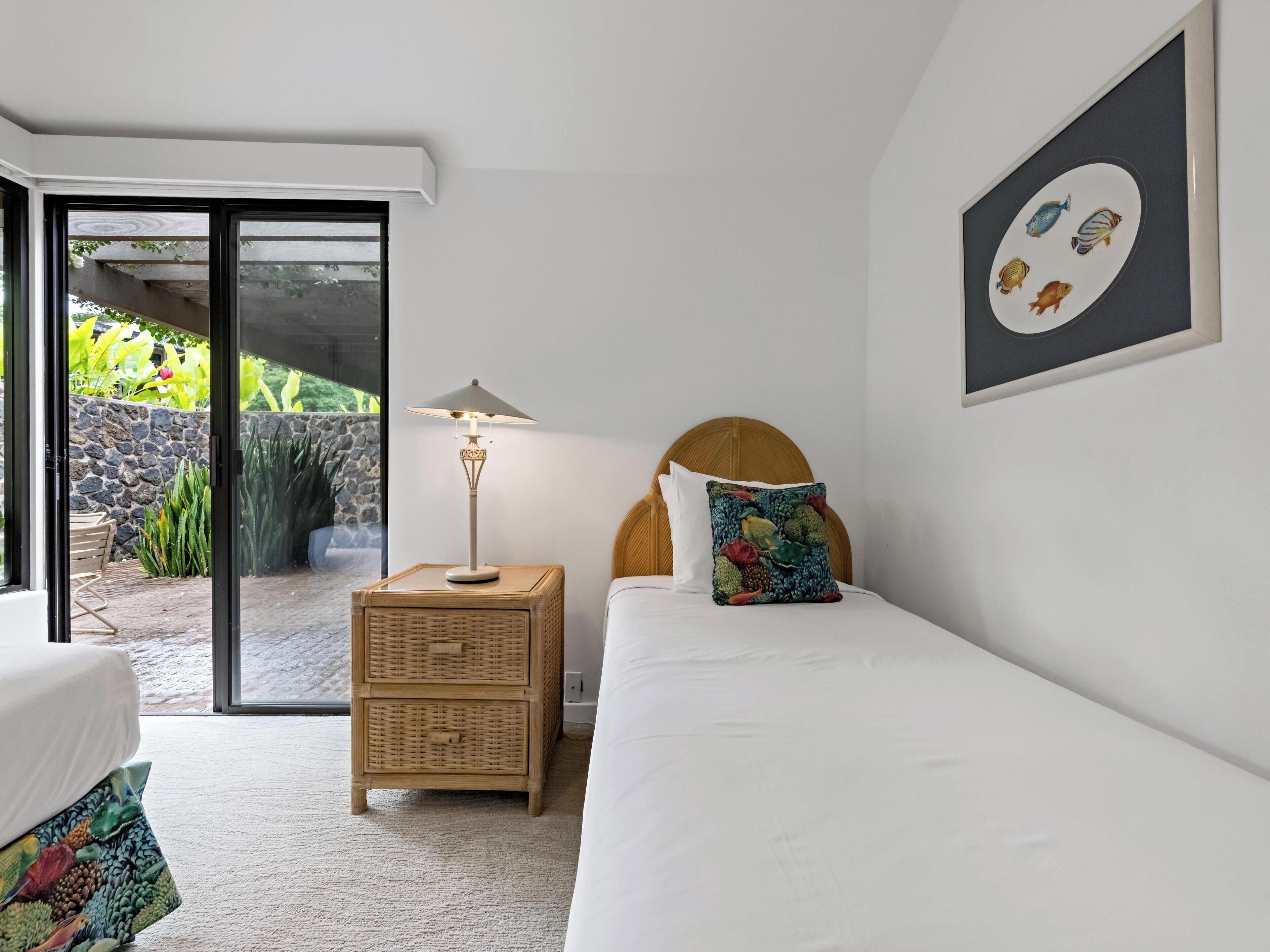 Quarto com cama, mesa de cabeceira com abajur, mesinha de palha e porta que dá para uma área externa, além de um quadro na parede.