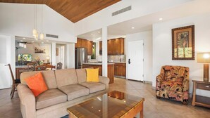 Ein geräumiges Wohnzimmer mit einer Couch, einem Couchtisch und einem gemusterten Sessel. Im Hintergrund ist eine Küche mit Holzschränken und einem Kühlschrank erkennbar.
