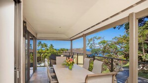 A balcony with a dining table and chairs, a vase with flowers, and a view of palm trees and the ocean.