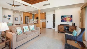 A spacious living area with a wooden ceiling, a sofa with patterned cushions, a wicker chair, a television, and a kitchen in the background.