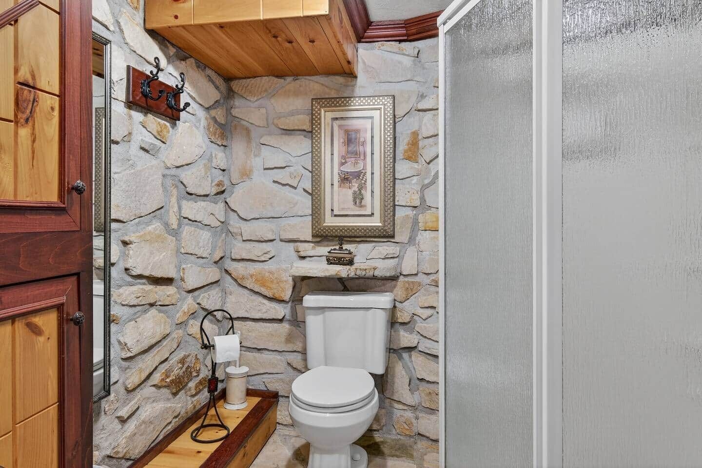 Ein Badezimmer mit Steinwand, Holztür, einem gerahmten Bild, einer Toilette und einer kleinen Ablage mit einer Flasche und einem Stecker.