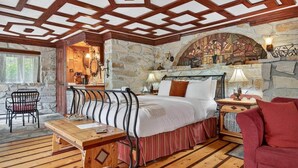 Une pièce chaleureuse avec un mur en pierre, un lit doté d’une tête de lit en bois, un fauteuil rouge, une table en bois et une cheminée en pierre ornée d’une fresque.