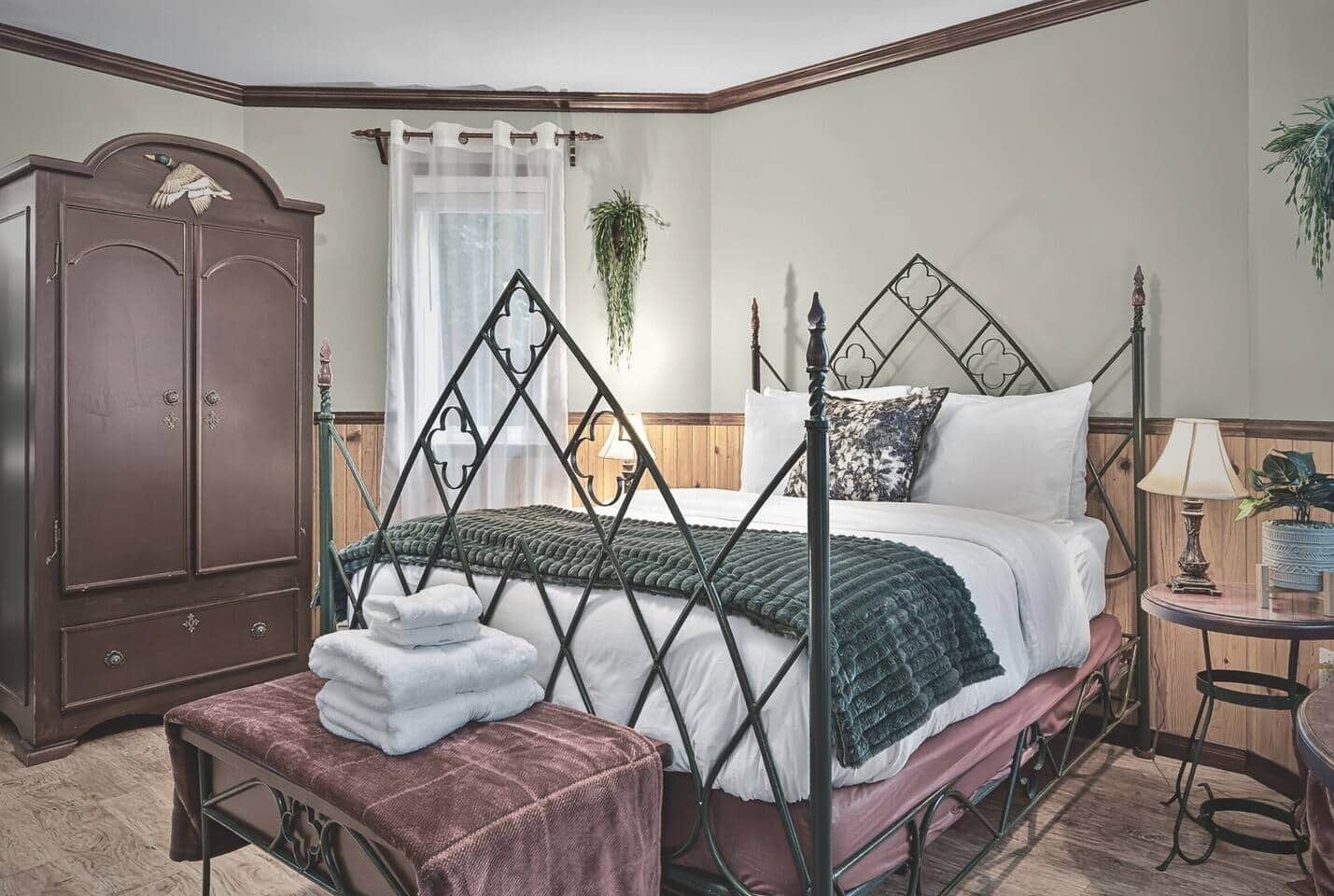 A bedroom with a four-poster bed, a wooden wardrobe, a bedside table with a lamp, and a hanging plant.
