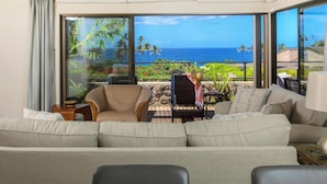 Un salon spacieux avec un grand canapé, un fauteuil et une table. Une grande fenêtre offre une vue sur l’océan et le paysage tropical.