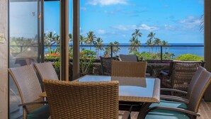 Un balcon doté de meubles en osier et d’une table en verre, offrant une vue sur une plage tropicale bordée de palmiers et sur l’océan.