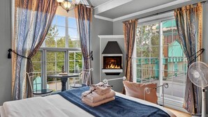 Une chambre avec une cheminée, un lit recouvert d’une couette bleue et une vue sur une maison verdoyante, visible à travers de grandes fenêtres.
