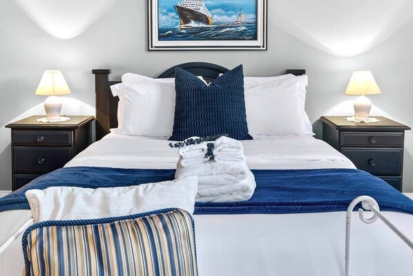 Un lit bien fait, recouvert d’une couverture bleue, avec des oreillers blancs et un coussin bleu foncé. Il y a deux tables de chevet avec des lampes, et un tableau représentant un navire est accroché au mur.