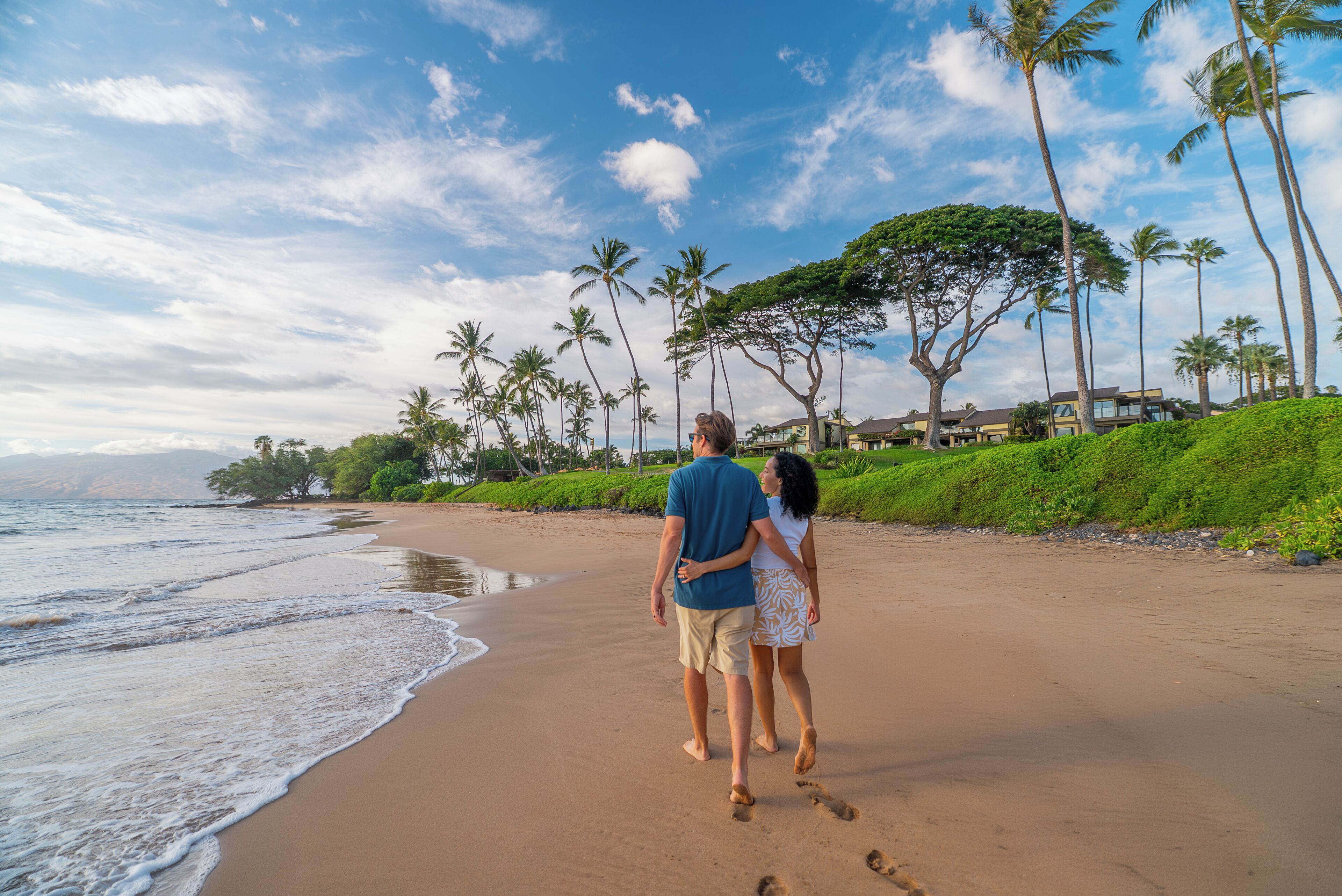 Un couple se promène sur une plage de sable, bordée de palmiers, sous un ciel dégagé.