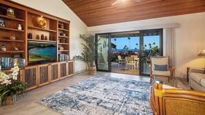 Een ruime woonkamer met een grote televisie, houten planken, een grote kamerplant en uitzicht op de oceaan door een schuifdeur.