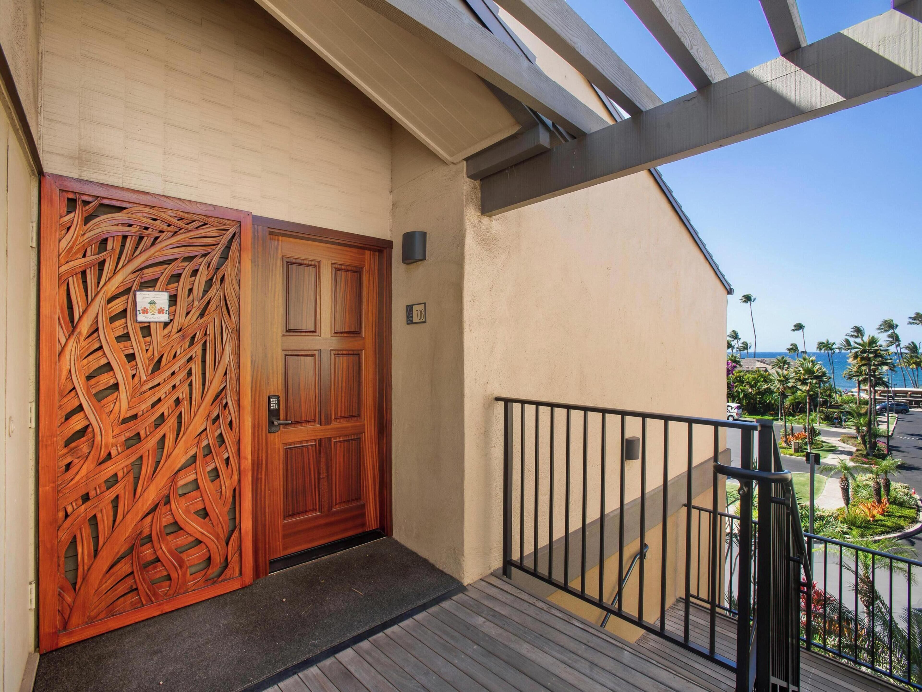 Una porta in legno con pannello decorativo, un balcone con ringhiera metallica e vista sull'oceano e palme.
