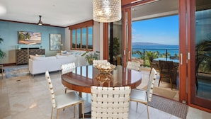 A spacious living room with a large round table, white woven chairs, and a view of the ocean through large sliding glass doors. - Modern Ocean View Elegance - Wailea Beach Villas PH404 (Wailea)