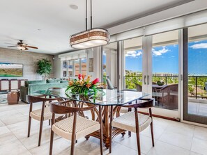 Un espace repas moderne avec une table en verre et des chaises en osier, un ventilateur de plafond et une vue sur un balcon aménagé avec des sièges d’extérieur.