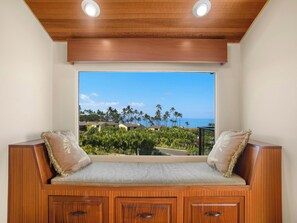 A wooden bench with cushioned seating and storage cabinets, overlooking a tropical landscape with palm trees and a view of the ocean. - Spacious Condo - Great for Families (Wailea)