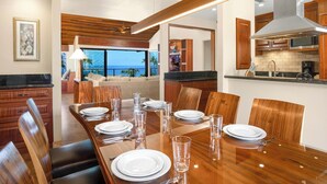 A modern dining area with a wooden table set for six, a kitchen with wooden cabinets, and a view of the ocean. - Spacious Condo - Great for Families (Wailea)