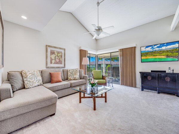A spacious living room with a sectional sofa, a glass coffee table, a ceiling fan, and a large TV on the wall. - Walk to Waves, Snorkeling, Pool - Wailea Elua 910 (Wailea)