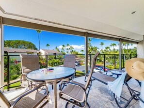 A covered outdoor seating area with a round table and chairs, overlooking a parking lot and greenery. - Walk to Waves, Snorkeling, Pool - Wailea Elua 910 (Wailea)