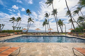 A swimming pool with lounge chairs and umbrellas, surrounded by palm trees and a view of the ocean. - Wailea Elua Village 910 | Remodeled, Vaulted Ceilings, 2 Min to Ulua Beach and P (Wailea)