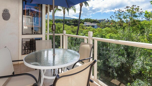 A balcony with a glass table and chairs, a blue umbrella, and a view of lush greenery and houses. - Quiet Retreat Between Fairways - Grand Champions 140 (Wailea)