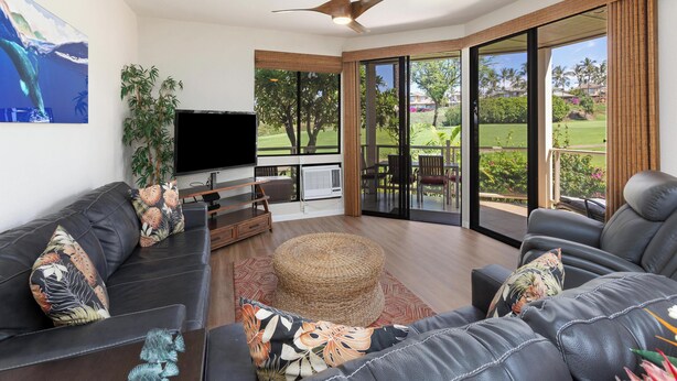 A modern living room with a flat-screen TV, leather sofas, a woven ottoman, and a large sliding glass door leading to an outdoor area with a view of a golf course. - Large Floorplan - Great for Families | Grand Champions 159 (Wailea)