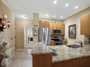 A kitchen with granite countertops, stainless steel appliances, wooden cabinets, and a decorative wall piece. - Golfer's Dream | Condo on Wailea Course (Wailea)