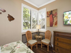 Une chambre à coucher avec un lit, deux fauteuils en osier, une petite table ronde avec une lampe, une commode, une fenêtre avec des stores et un tableau accroché au mur.