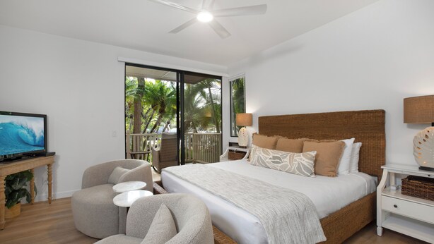 Une chambre avec un lit, une télévision, un ventilateur de plafond, un balcon donnant sur des palmiers et une tête de lit en osier.