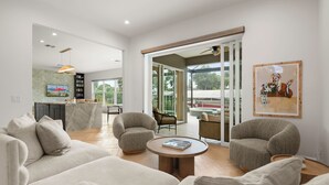 Una sala de estar moderna con un sofá, sillones y una mesa de centro. Se aprecia una cocina al fondo con una mesa de comedor y sillas.