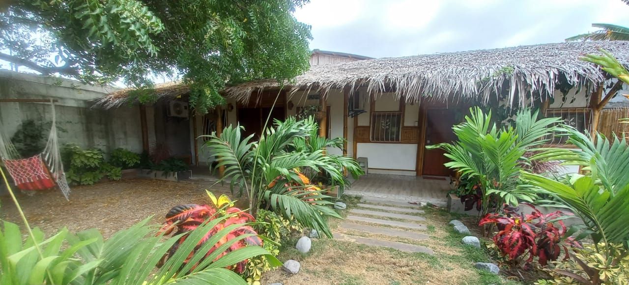 Una casa con techo de paja, rodeada por un jardín con diversas plantas y una hamaca.