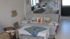A living room with a sofa, a coffee table, and a dining table. There is a kitchen in the background with appliances like a microwave and a refrigerator. - Pen Ty lumineux avec jardinet clos, en plein cœur de la côte de granit rose (Trégastel)