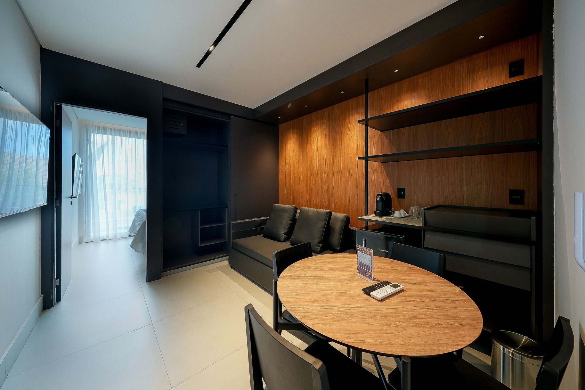 A modern living room with a round wooden table, black leather sofa, and built-in wooden shelving.