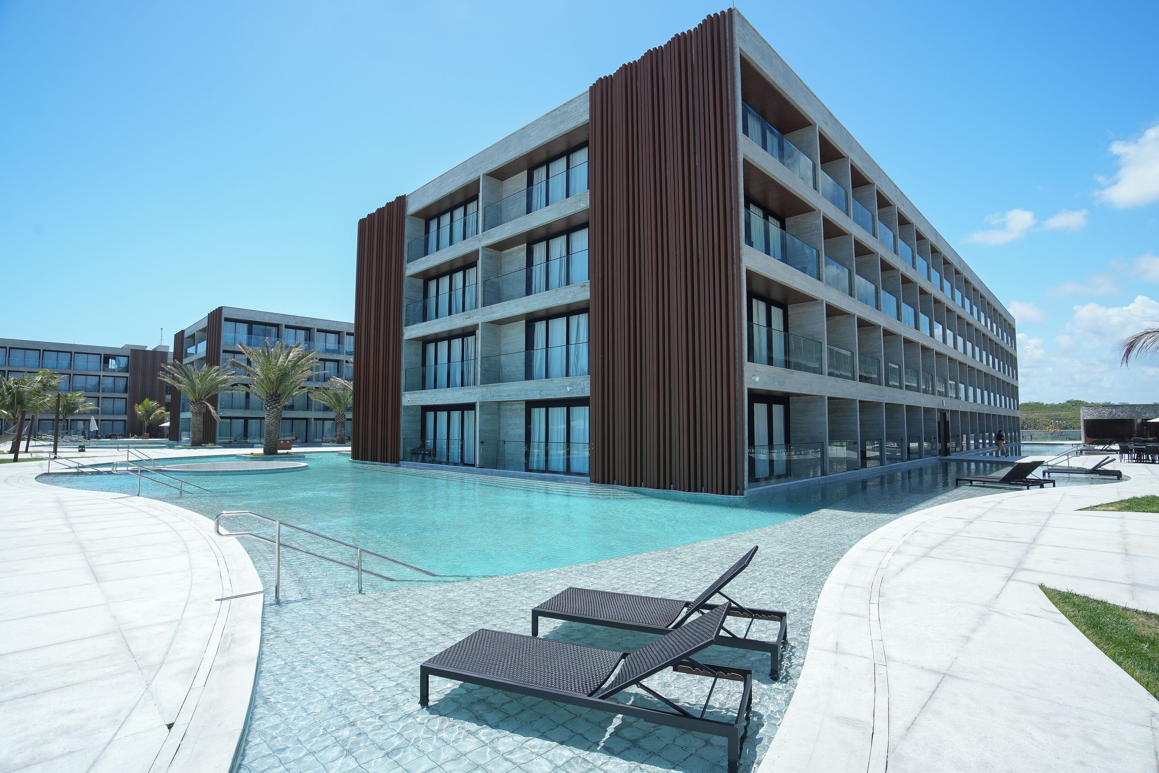 A modern building with a swimming pool, lounge chairs, and palm trees.