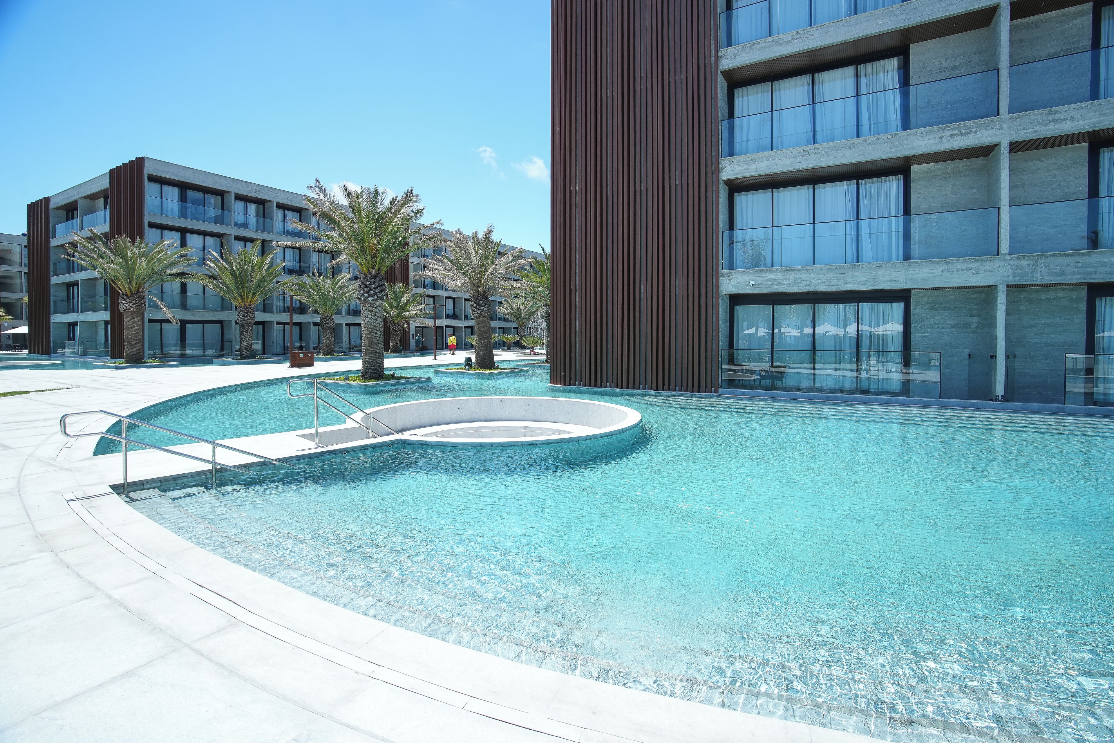 A modern hotel with a swimming pool, palm trees, and a clear blue sky.