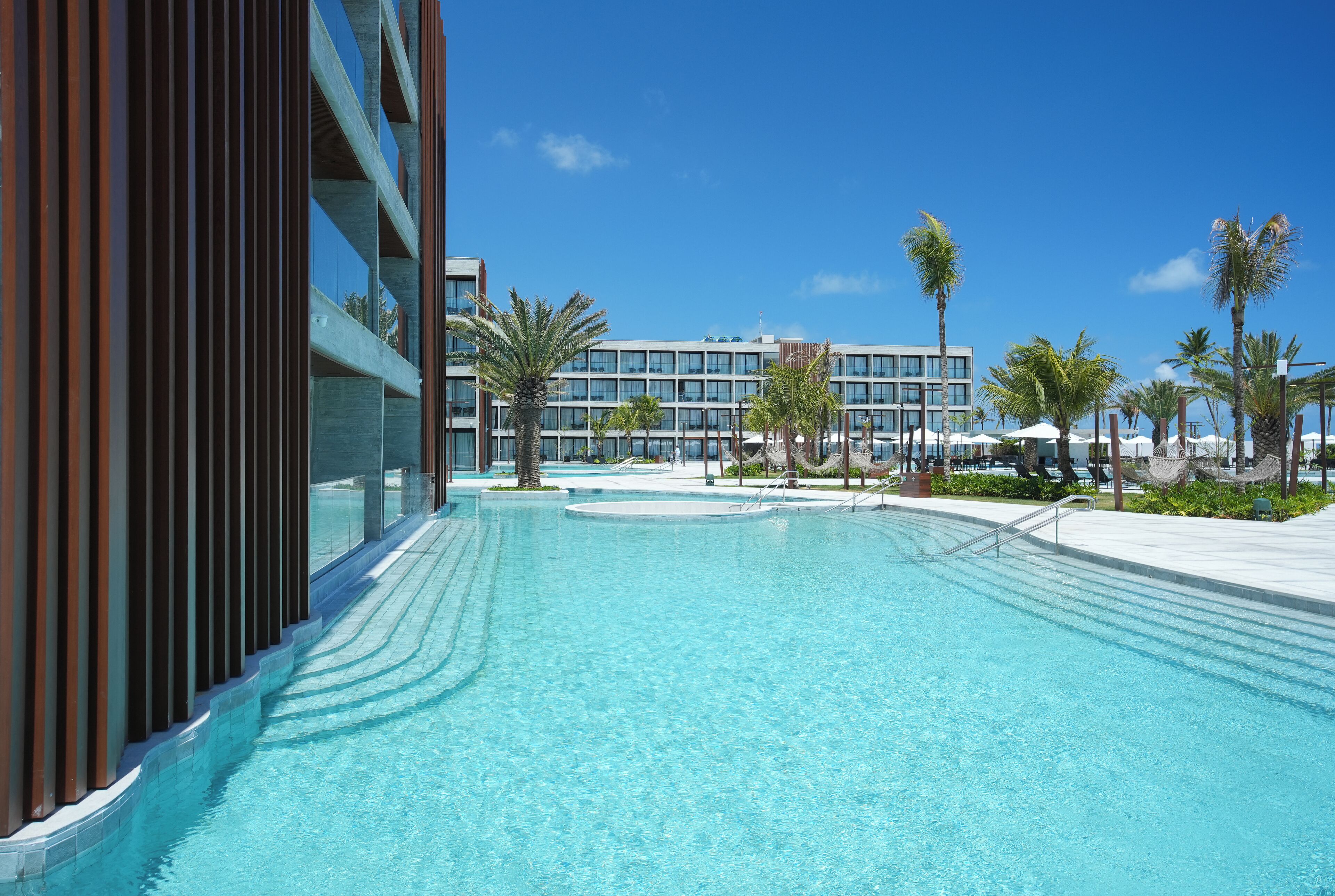 Une piscine à l’eau cristalline, entourée de palmiers et bordée d’un bâtiment moderne à plusieurs étages.