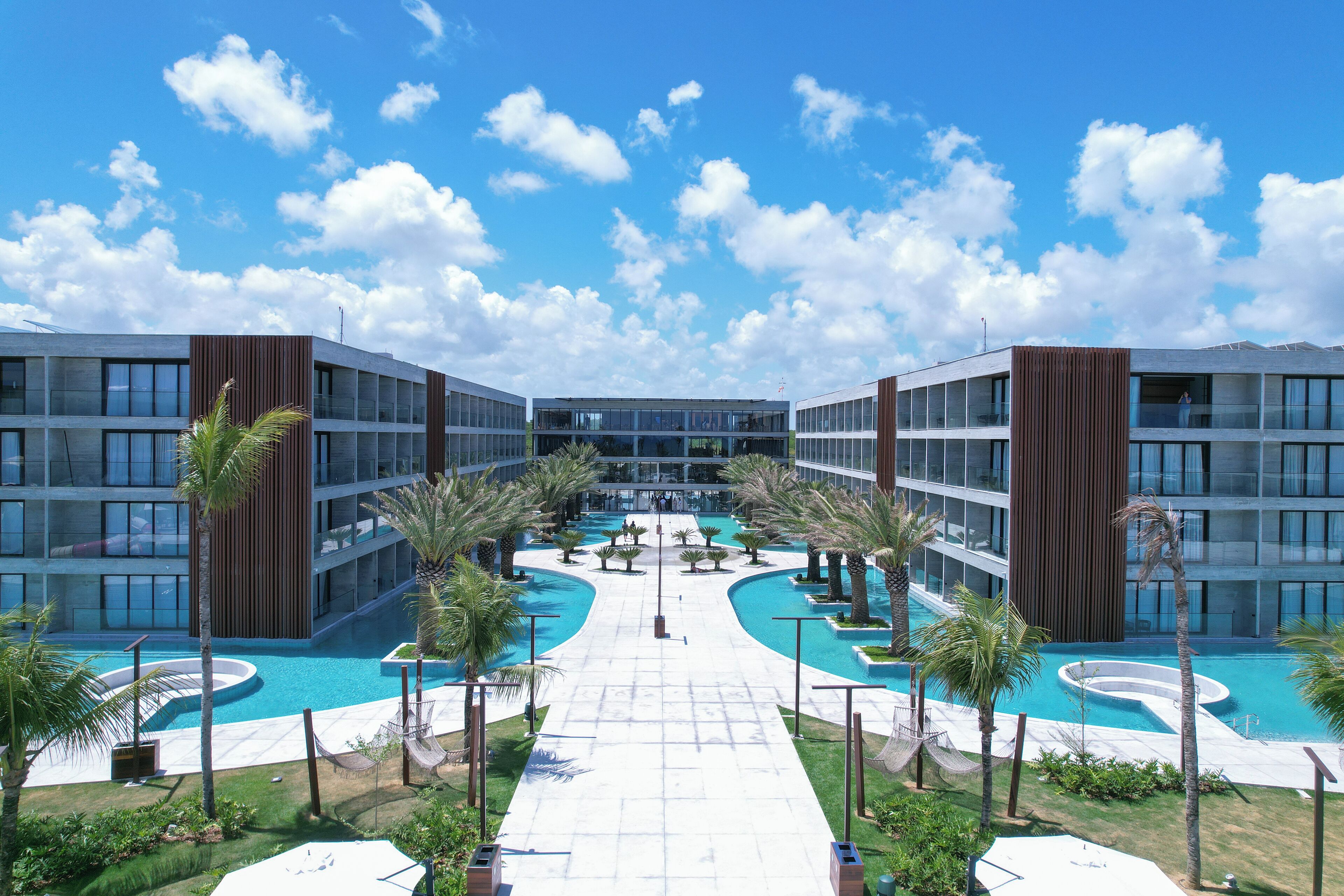 A modern hotel with a central swimming pool, surrounded by palm trees and a clear blue sky.