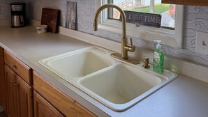 A kitchen with wooden cabinets, a double sink, a coffee maker, and a window with a view of trees. - Lake it Easy 2-bedroom cottage in Lyndonville (Lyndonville)