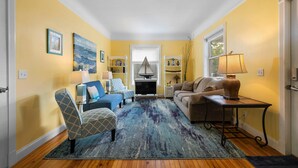 Un salon avec un fauteuil à motifs bleus, un canapé beige et une table basse. On y trouve un téléviseur posé sur un meuble, une lampe et une bibliothèque agrémentée d’objets décoratifs.