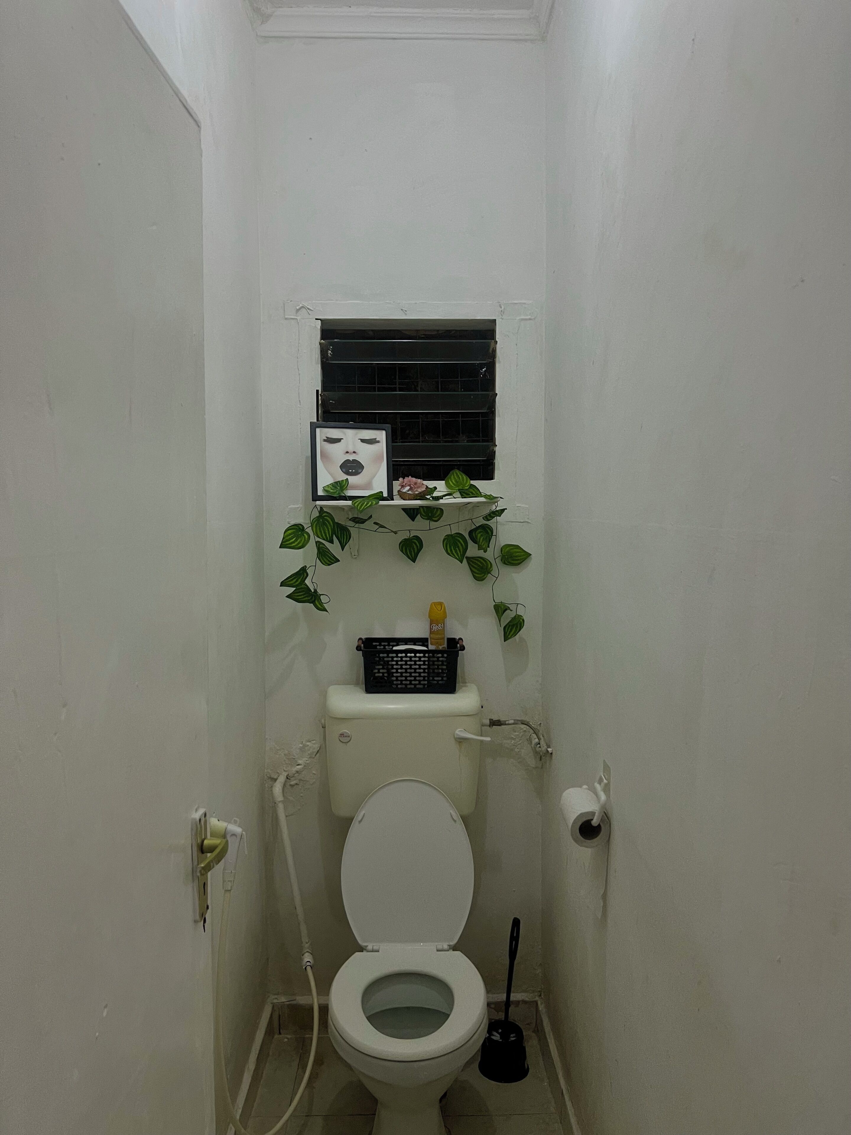 A small, white bathroom with a toilet, a window above the toilet, and a small shelf with a picture frame and a bottle.