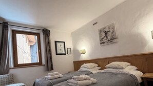 A bedroom with two beds, a wooden headboard, a small table, a bench, and a window with curtains. - Apartment in the old village, at the foot of the slopes. Residence le SQUAW VALLEY (Val-d'Isère)