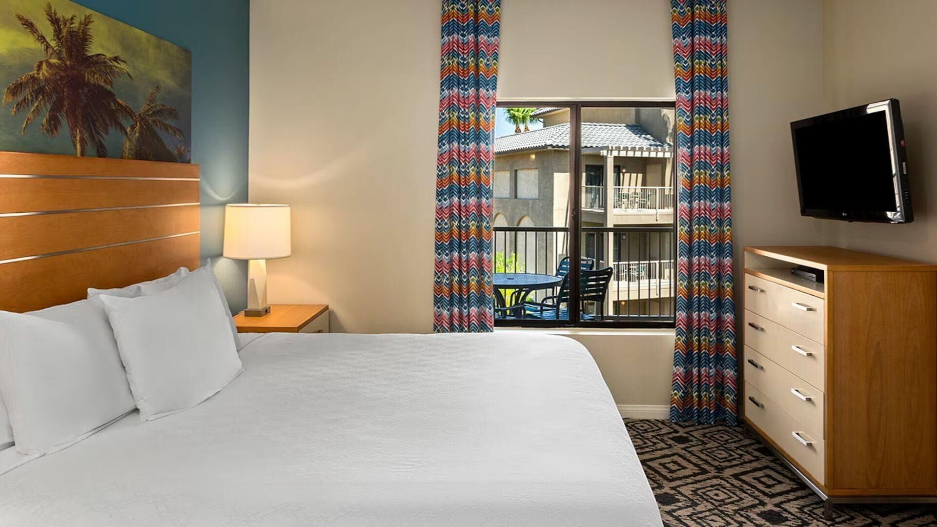 Una habitación de hotel con una cama, una mesita de noche con una lámpara, un televisor montado en la pared, una ventana con cortinas de colores y vistas al balcón exterior.