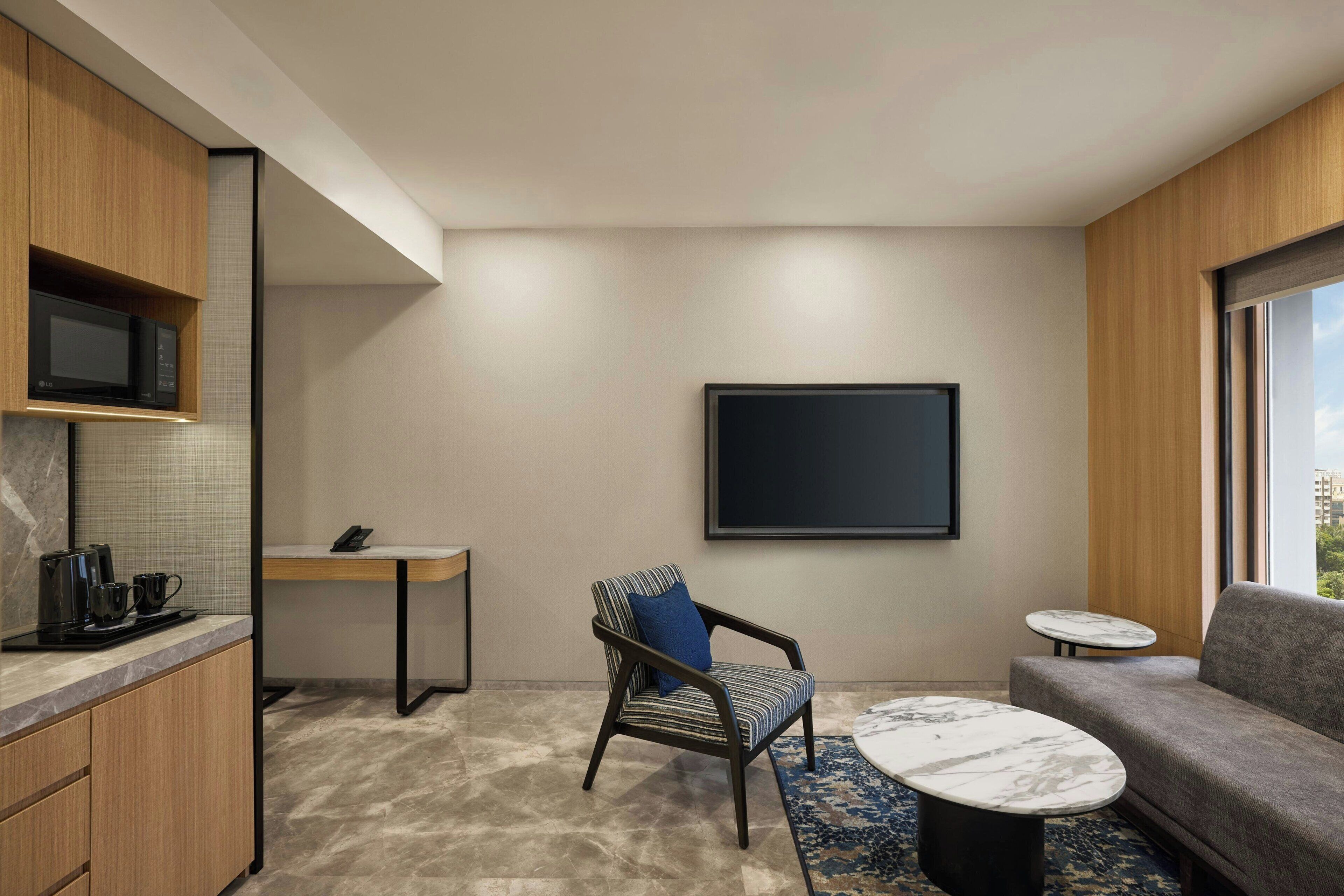 Une chambre d’hôtel moderne dotée d’un coin cuisine, d’un espace salon avec un canapé et un fauteuil, d’une petite table ronde et d’une grande fenêtre donnant sur la ville.