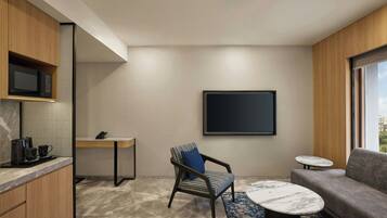 Une chambre d’hôtel moderne dotée d’un coin cuisine, d’un espace salon avec un canapé et un fauteuil, d’une petite table ronde et d’une grande fenêtre donnant sur la ville.