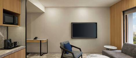 Una habitación de hotel moderna con cocina, zona de estar con sofá y sillón, una pequeña mesa redonda y un amplio ventanal con vistas a la ciudad.