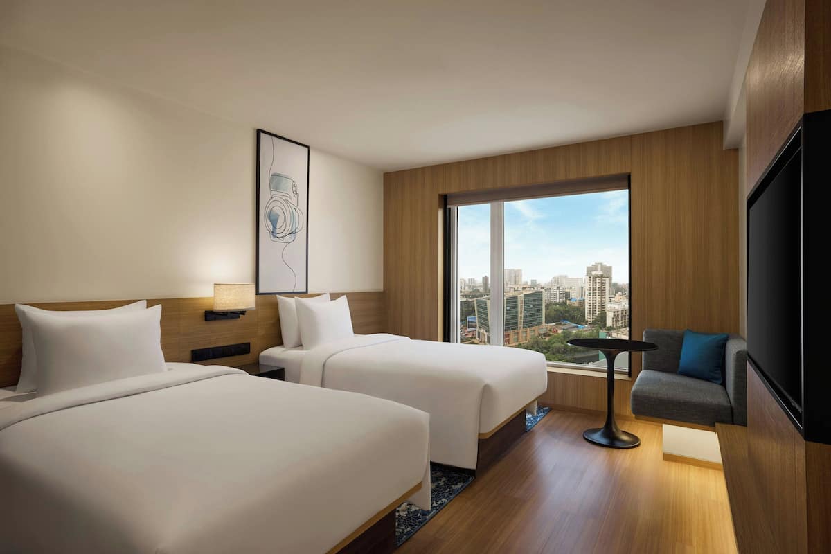 Habitació d'hotel amb dos llits, un sofà, una televisió, una finestra gran amb vista a la ciutat i una obra d'art emmarcada a la paret.