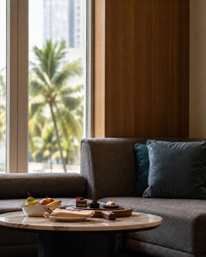 A modern living room with a sofa, a round table with food, and a view of palm trees through large windows.