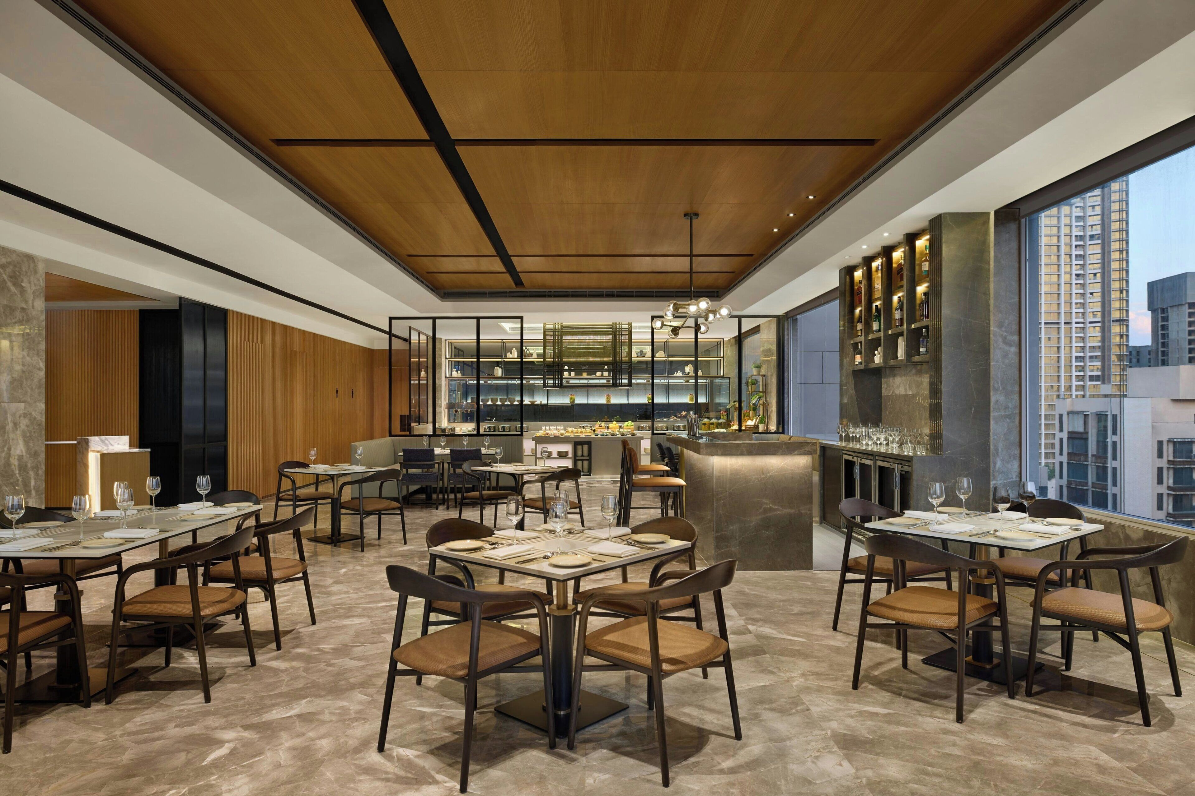 Un restaurante moderno con suelos de mármol, techo de madera y amplias ventanas con vistas a la ciudad. Las mesas están dispuestas con copas de vino y platos.