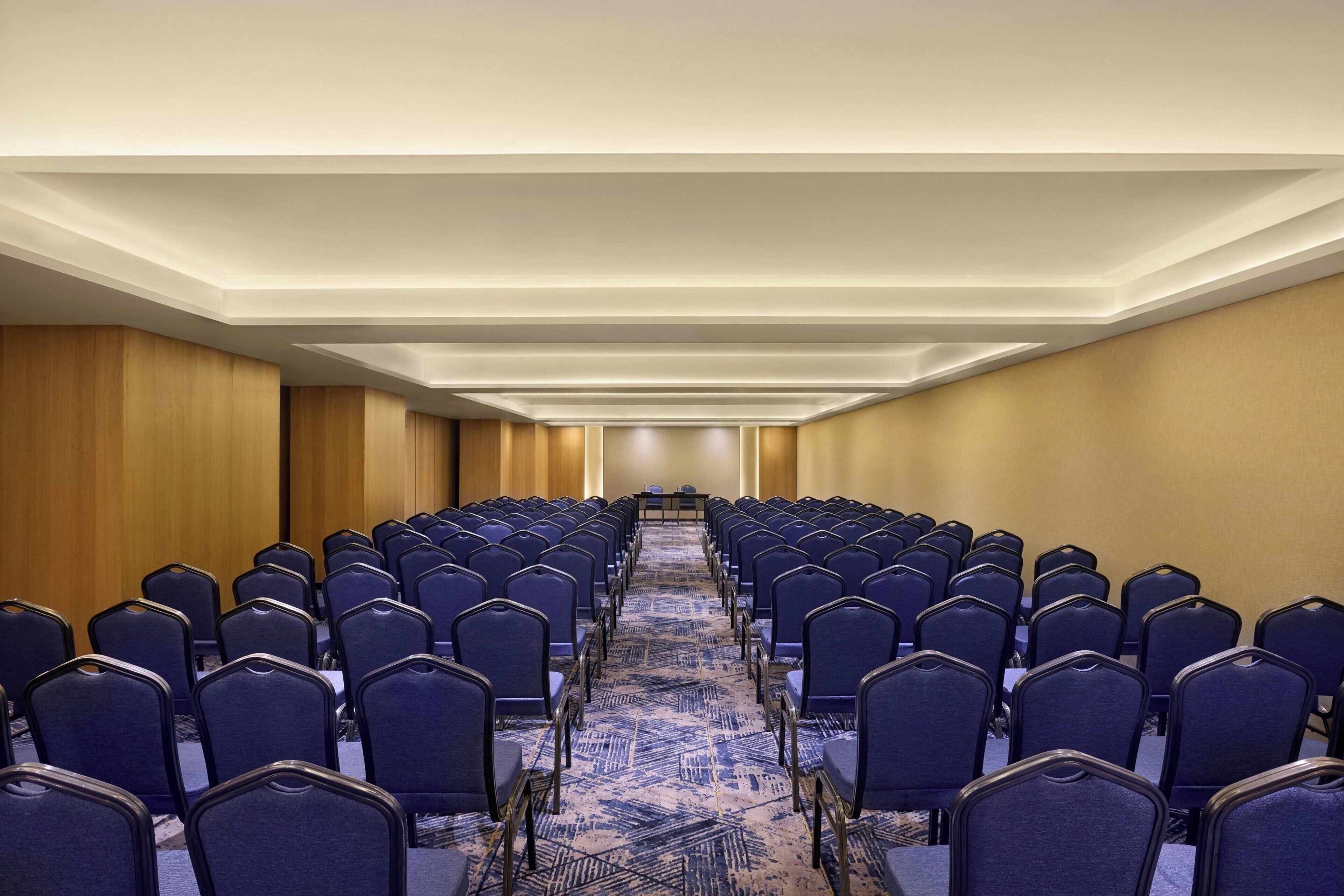 A conference room with rows of blue chairs, a long table at the front, and a patterned carpet.