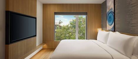 A modern hotel room with a large bed, a flat-screen TV mounted on the wall, and a large window offering a view of the outdoors.
