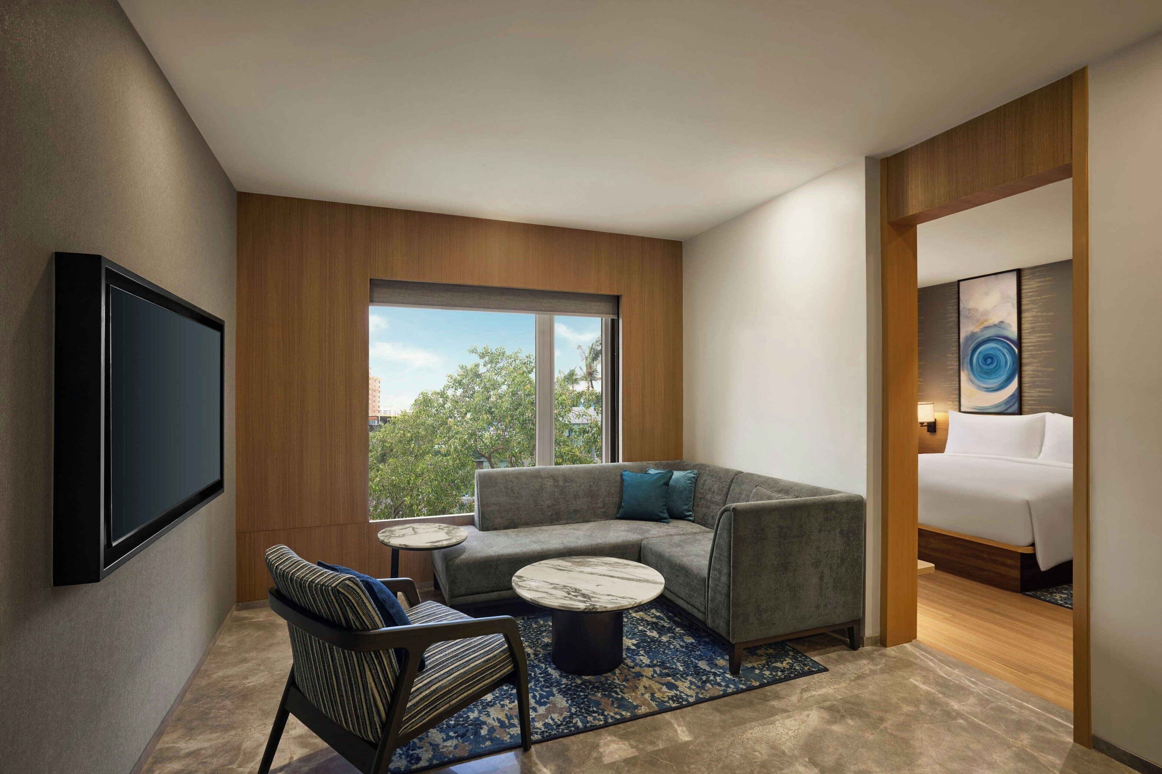 A modern hotel room with a large window, a sofa, a chair, a small round table, and a bed.